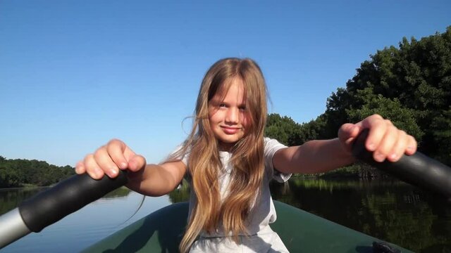 Little smiling girl rowing along the river and looking at camera. Child controls the boat independently. Kid sails on a boat outdoors on summer vacation. Fishing with dad