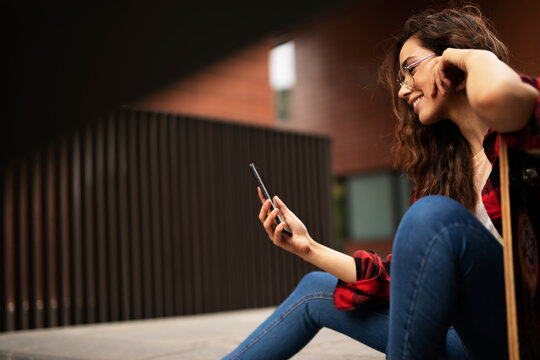 Portrait of young beautiful girl with skateboard. Happy smiling woman using the phone