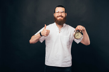Young man wearing glasses is holding an alarm clock and showing thumb up.