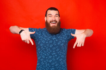 Wide smiling bearded male hipster is pointing down with both hands.