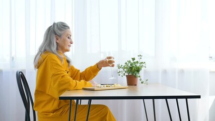 Beautiful old grandmother with grey hair and face with wrinkles sitting at the table at home on window background with glass of champagne, mother's day, happy retirement