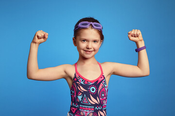 Little girl spends leisure time in water park, raises arms and shows muscles, ready for swim, poses...