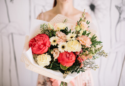 Very nice young woman holding big and beautiful bouquet of fresh gerbera daisy, coral peony, carnations, eustoma, pistachio branches flowers in coral and yellow colors, cropped photo, bouquet close up