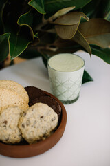 Delicious green Matcha latte in a glass standing on the white table with a plate full of cookies and a vase with Magnolia leaves