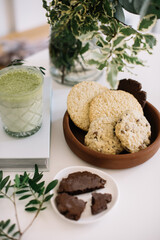 Delicious green Matcha latte with a plate full of cookies and another plate with one crumbled chocolate cookie, all standing on the white table with some greenery in a vase