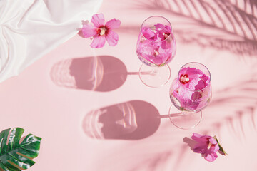 Creative composition made of two glasses with hibiscus flower water on pastel pink background with palm tree leaves shadows and monstera leaf. Summer and refreshment concept