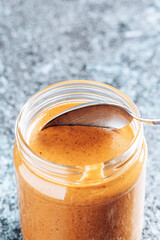 Spoon dipping into a glass jar with crunchy peanut butter. Place for text. Food background.