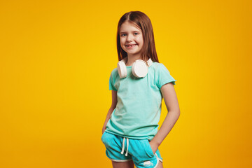 Confident girl in stylish summer outfit, wearing white headphones on neck, keeping hands in pockets, isolated over yellow brick wall