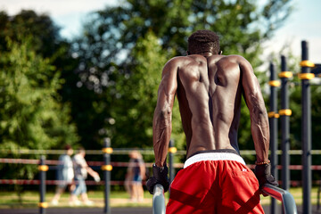 Muscular african american athlete in with a naked sculpted torso doing push-ups on the uneven bars in the urban landscape, Back shot, copy space