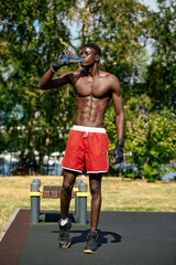 Young african american man drinks water while training in the park on the sports ground, outdoor workout concept, crossfit