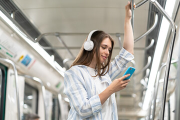 Young girl listen music in headphones and hold smartphone in subway car travel underground use...