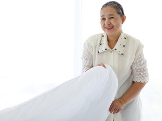 Happy smiling Asian senior elderly woman housewife cleaning and making bed at bedroom, grandma doing housework and cleaning, old female chambermaid changing the white blanket and sheet