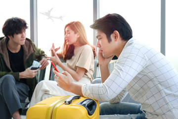 Asian young man holding and looking at mobile phone while waiting airline flight to travel, showing...