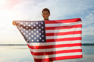 Young African American man looking at camera and proudly holding American flag on blue sky background outdoors near river at sunset