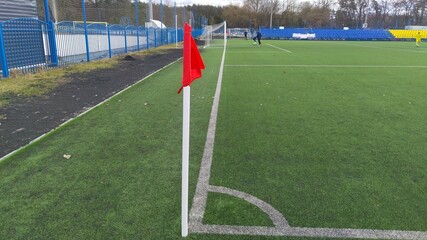 The corner flag is placed on the artificial football field of the city stadium with metal and mesh fences and grandstand for fans