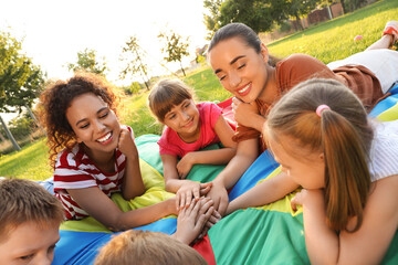 Group of children with teachers holding hands together on rainbow playground parachute in park. Summer camp activity