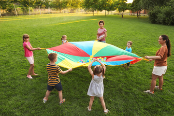 Group of children and teachers playing with rainbow playground parachute on green grass. Summer camp activity - obrazy, fototapety, plakaty