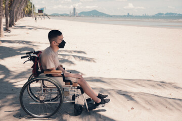 Disabled teenager boy wearing face mask on outdoors public place like everyone else, The sea beach background, Lifestyle of handicapped vacation, Diverse people concept.