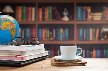 White coffee cup, eyeglasses with books stacked and black laptop on wooden table with blurred bookshelf in home office room area 