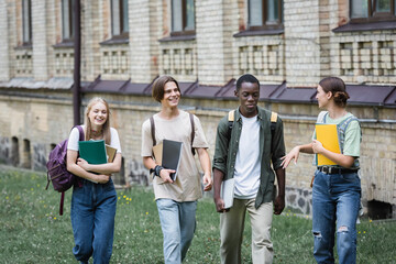 Smiling interracial students with laptop and notebooks walking near friend outdoors