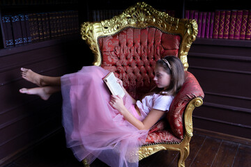 Lovely teen girl in pink tutu-skirt reading book while sitting sideways on chic gold-lined armchair