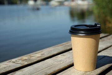 Obraz premium Takeaway cardboard coffee cup with plastic lid on wooden pier near river, space for text
