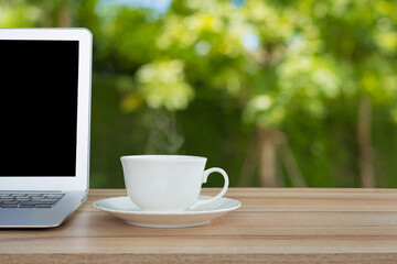 A white coffee mug and laptop is placed on old wooden top table with blurred green leaves, bokeh in garden background. Good morning or have a happy day and positive message concept.