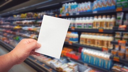 Hand with note paper at supermarket showcase background. Shopping list with copy space. Selective focus on paper