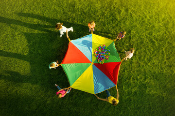 Group of children and teachers playing with rainbow playground parachute on green grass, top view....