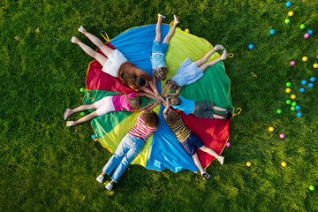 Group of children with teachers holding hands together on rainbow playground parachute in park, top...