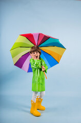 a funny little boy in a green raincoat and yellow rubber boots stands with a multicolored umbrella stands on a blue background with a place for text