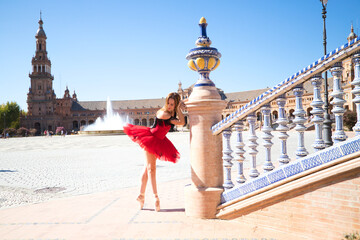 ballet dancer with red tutu leaning on a park railing in seville. The dancer makes different postures and stretches on the railing. Classical ballet concept.