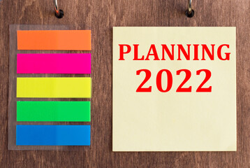 Text PLANNING 2022 on yellow paper on a wooden background next to colored stickers for notes