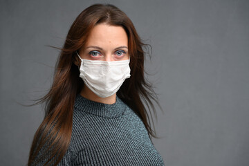Close-up portrait of adult brunette woman in medical mask on gray background. Model looks directly into the camera