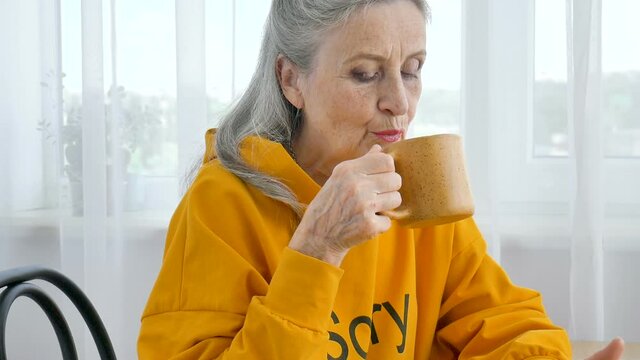 Happy elderly pensioner, eccentric gray-haired woman in yellow is drinking tea, looking thoughtfully into a mug