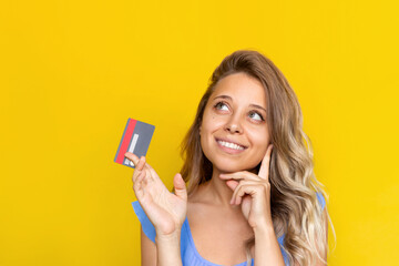 Close-up of a young blonde woman with wavy hair holding a plastic credit card in her hand thinking about where to spend the money looking up at empty copy space isolated on a color yellow background