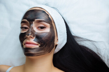 cosmetologist applying black mask on pretty woman face wearing black gloves, gorgeous woman in spa...