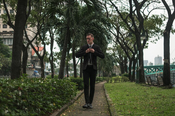 Attractive young caucasian man in black formal suit walking in the park. Park with tropical plants. Serious look. High quality photo