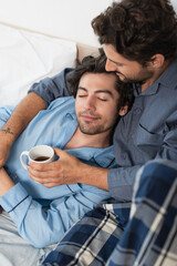 tattooed gay man holding cup of tea and hugging boyfriend in bed