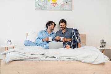 happy gay men watching movie on laptop and holding cups of tea