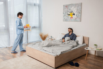 man holding breakfast tray with orange juice near boyfriend in bed