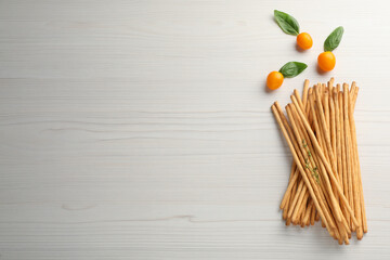 Delicious grissini sticks, basil leaves and yellow tomatoes on white wooden table, flat lay. Space for text