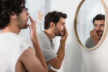 man applying cream on face near blurred boyfriend in bathroom