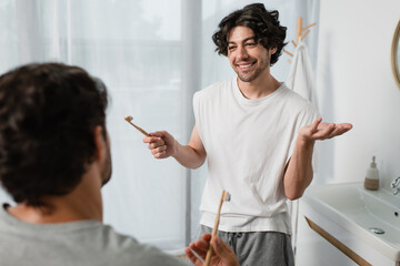 bearded gay man holding toothbrush while talking with blurred boyfriend in bathroom - Powered by Adobe