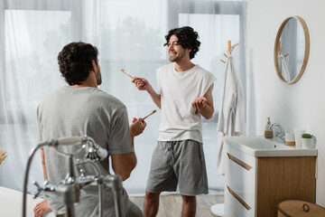 Fototapeta premium bearded gay couple holding toothbrushes while talking in bathroom