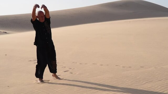 Old man in black is doing in kung fu in the dunes at sunset. a lot of sand, golden sunset light. sports training. handsome man meditating and dancing. Strong wind is blossing