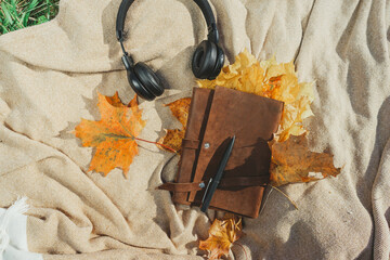 Top view of a leather book, diary and black headphones in autumn leaves lying on a blanket.