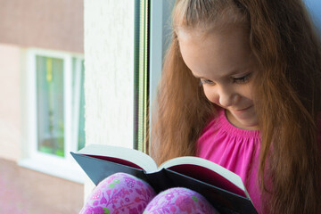 a sweet, dark-skinned little girl in a pink T-shirt and shorts sits at home alone in quarantine on the window sill in the summer and reads a book with interest. Lok Down education. copy space