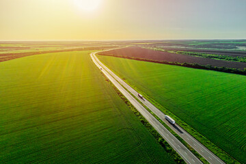 two white trucks driving on asphalt road along the green fields at sunset. seen from the air....