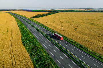 red truck driving on asphalt road aalong the yellow fields of wheat at sunset. seen from the air. Aerial view landscape. drone photography. cargo delivery